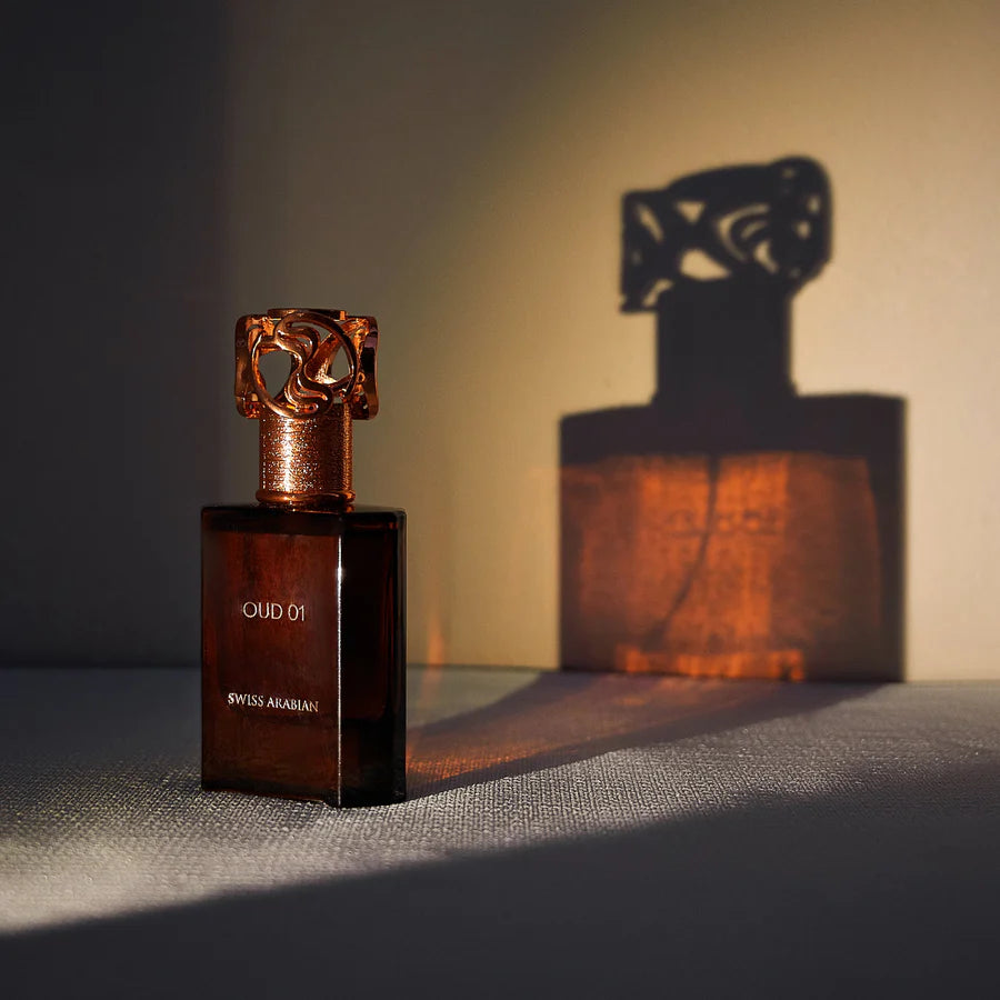 En mörk parfymflaska med texten 'OUD 01' och 'SWISS ARABIAN', stående mot en vägg där dess skugga projiceras dramatiskt i varmt ljus